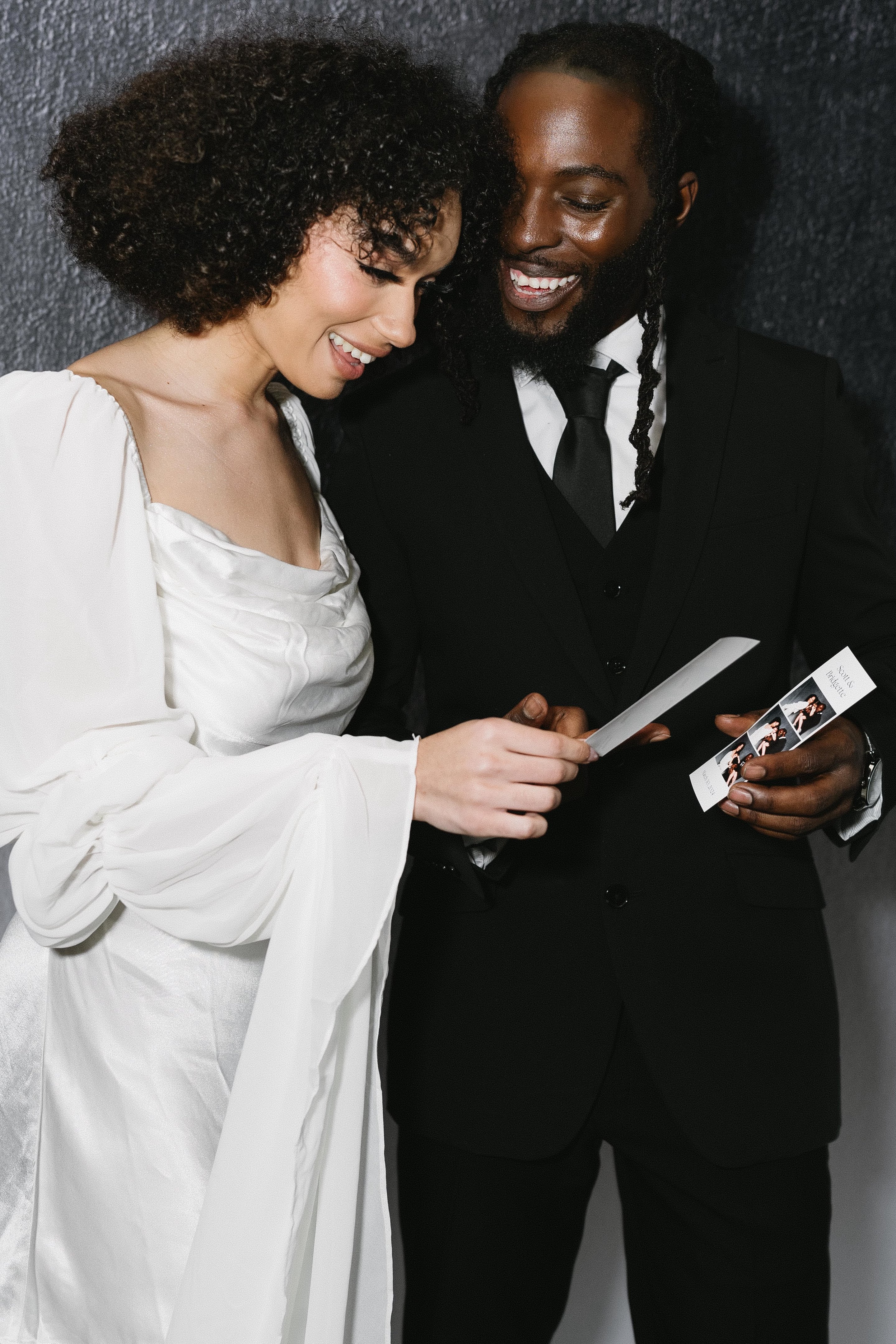 A couple in a white gown and black tie smiling over a strip of photo booth prints