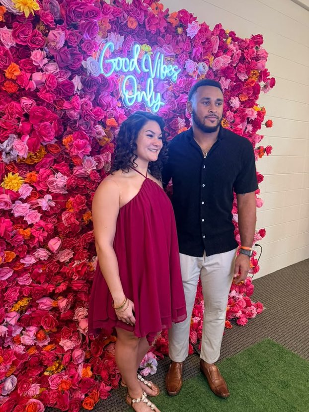 Two people posing by a pink flower wall with neon sign reading 'Good Vibes Only'